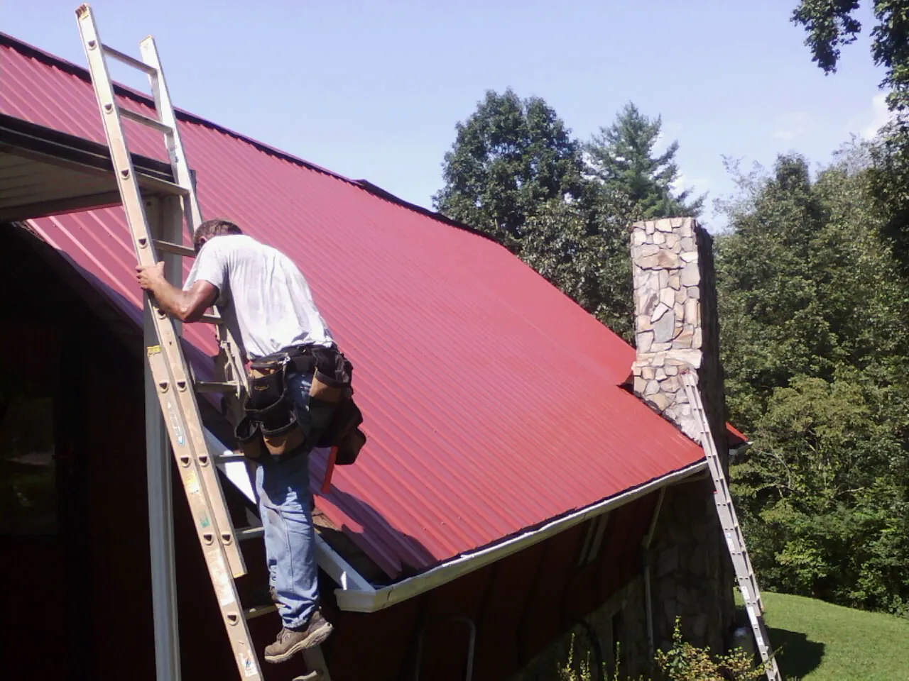 Roofer on a ladder beside a red metal roof and stone chimney — Soffit & Fascia Repair by Roofing Brothers in Beaver Dam